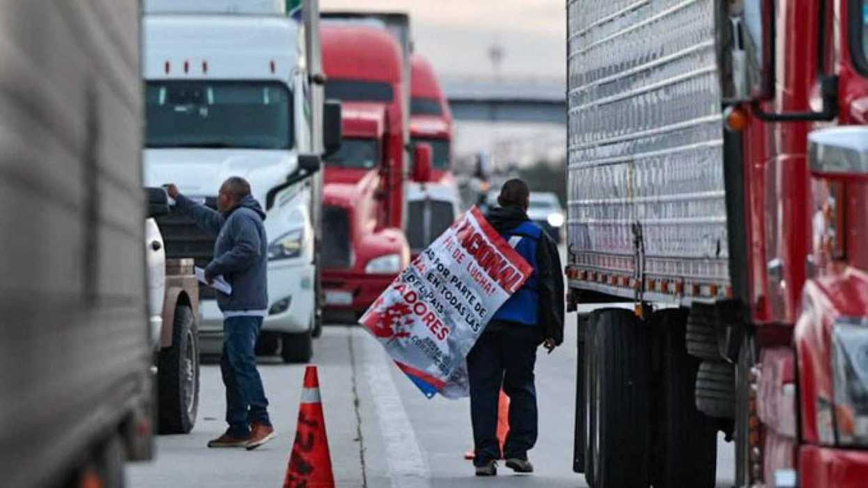 Truck drivers protest, holding a sign among parked semis on a busy road.
