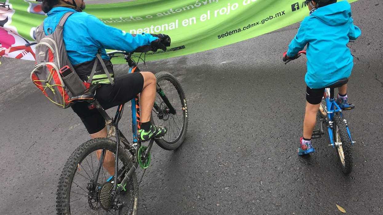 Two cyclists in blue jackets approach a banner on a wet street.