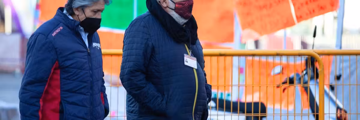 Two men in jackets and masks walking past colorful signs on a street.