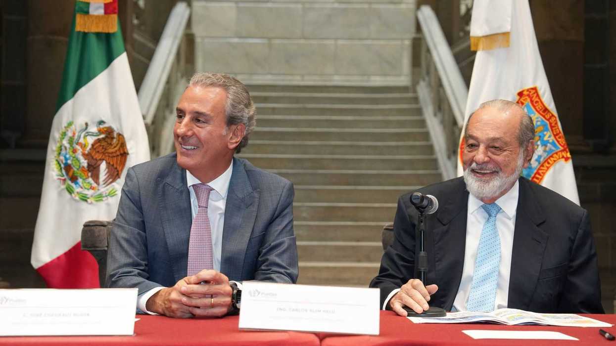 Two men in suits seated at a table with Mexican flags behind them.
