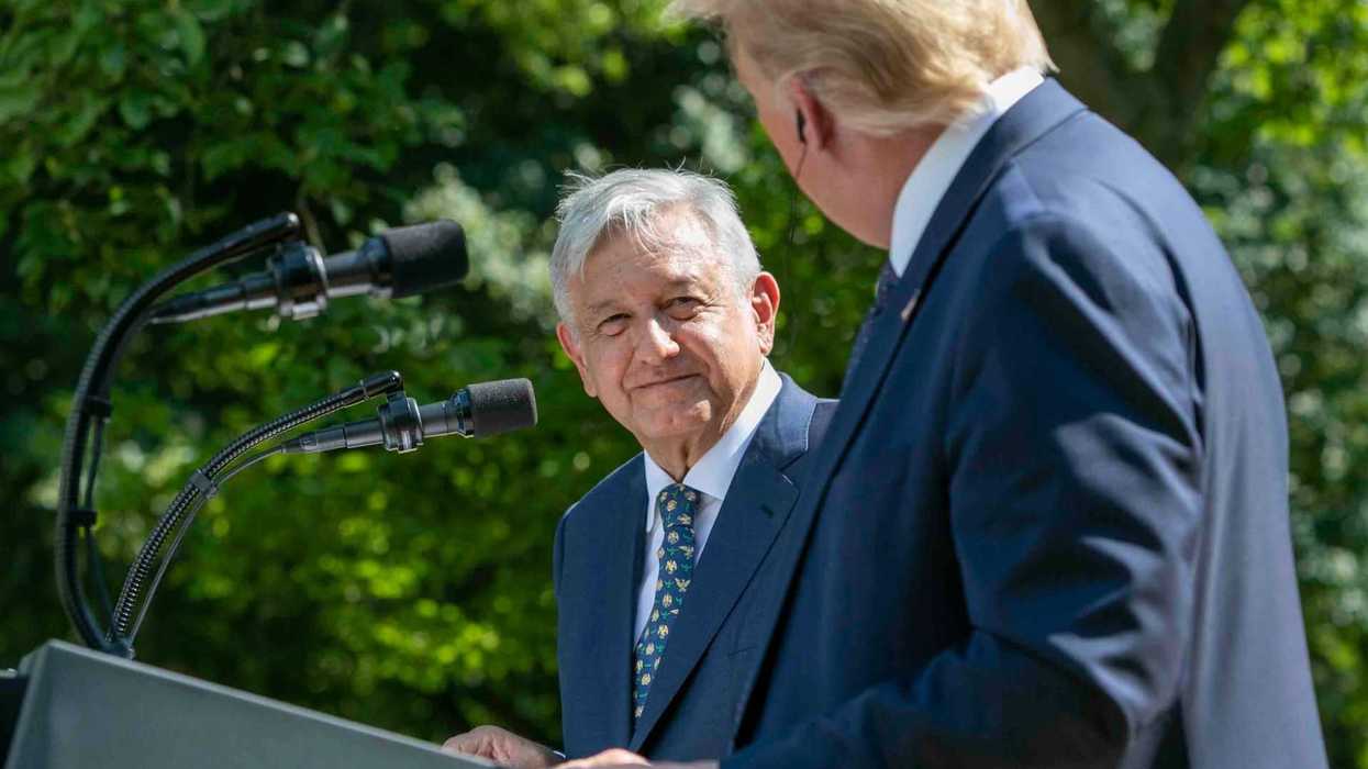 Two men in suits talk at podiums with trees in the background.