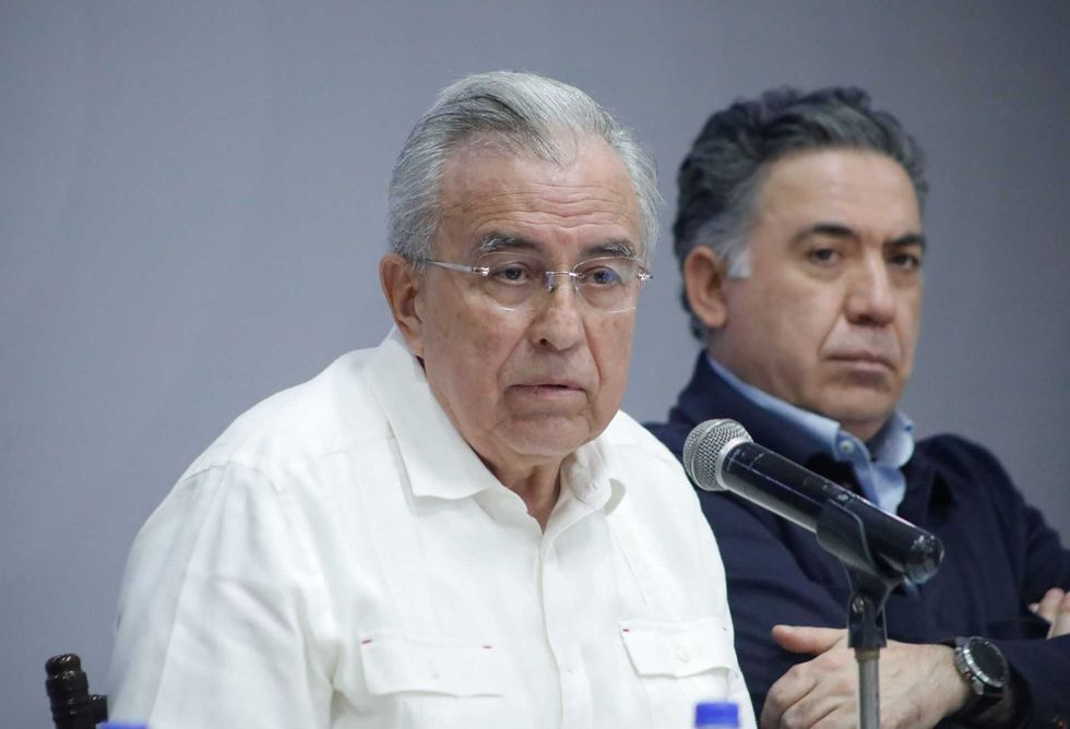 Two men sitting at a table, one speaking into a microphone.