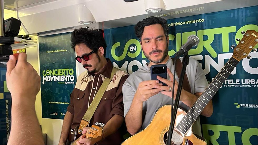 Two musicians stand with guitars at a radio studio, as one checks his phone beside a microphone.