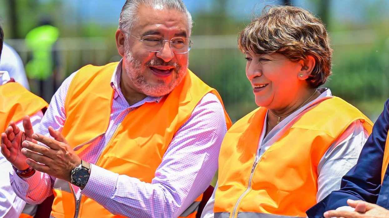Two people in orange vests smile and converse outdoors on a sunny day.