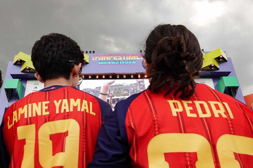 Two people in red soccer jerseys at a fan festival with a large screen.