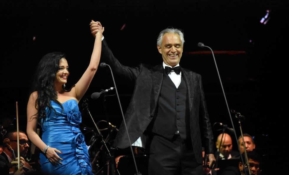 Two performers holding hands on stage, smiling, with an orchestra in the background.