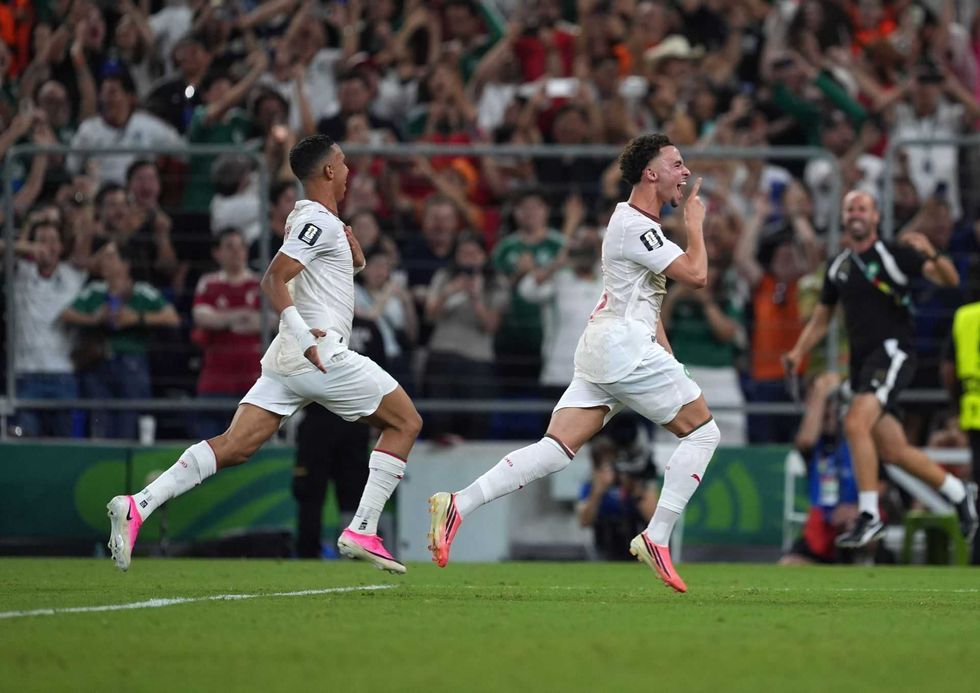 Two soccer players in white celebrate a goal; excited crowd in the background.