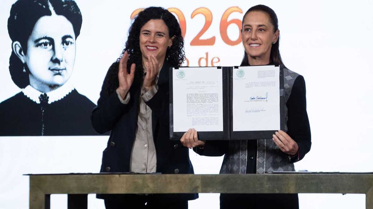 Two women at an event, one holds a signed document, portrait of a woman in the background.