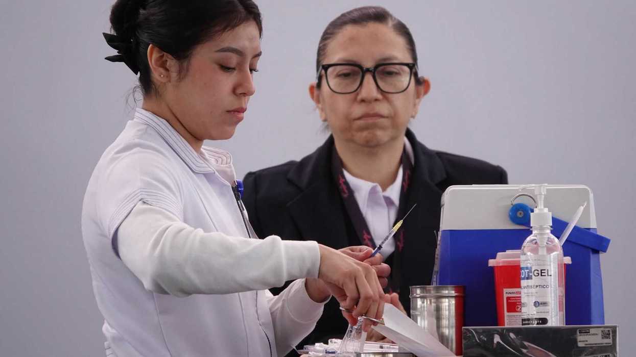 Two women in medical attire preparing a syringe at a workstation.