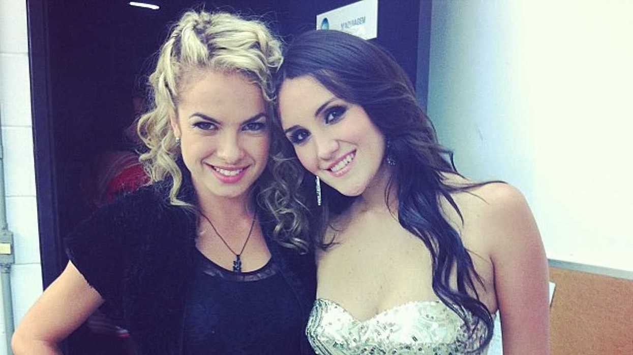 Two women smiling, one with curly hair, one with long dark hair, posing together indoors.