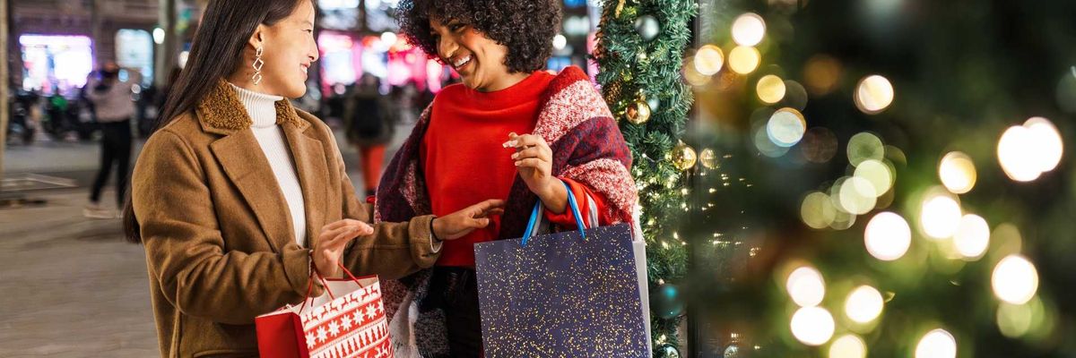 Two women smiling with shopping bags near festive holiday lights and decorations.