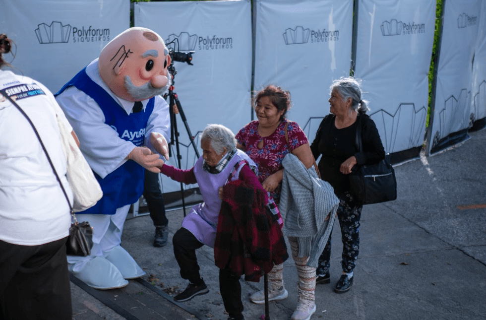 Una fotograf\u00eda de plano medio muestra a la mascota del Dr. Simi, con su tradicional bata blanca y bigote, saludando calurosamente a una se\u00f1ora