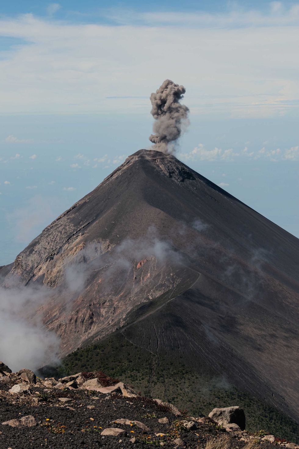Volcano emitting dark smoke against a cloudy blue sky.