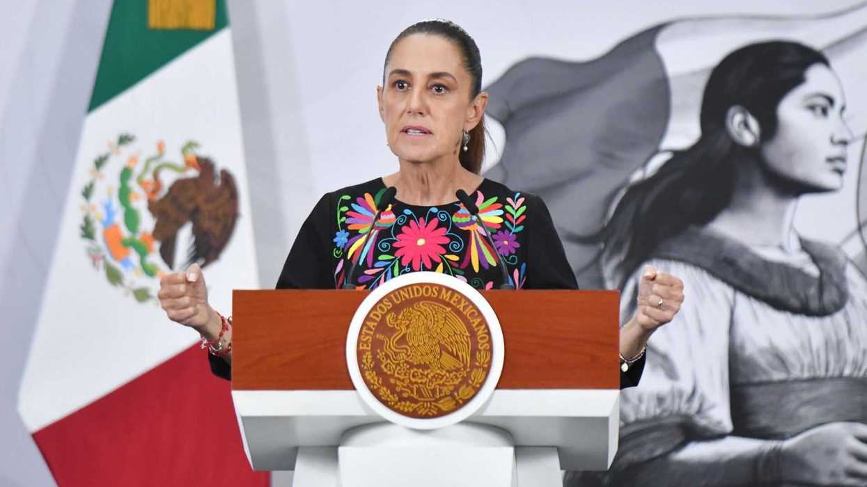Woman at podium with Mexican flag backdrop, wearing colorful floral blouse.