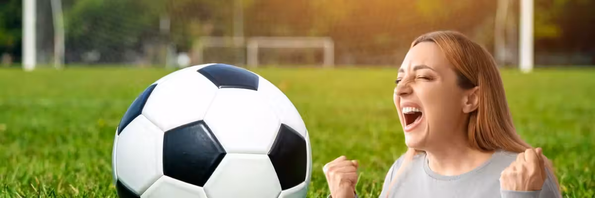 Woman cheering near a soccer ball on a grassy field with a goalpost in the background.