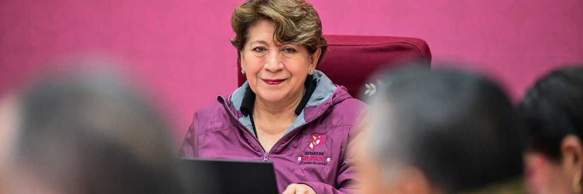 Woman in a magenta jacket smiles, seated at a table with others, against pink background.