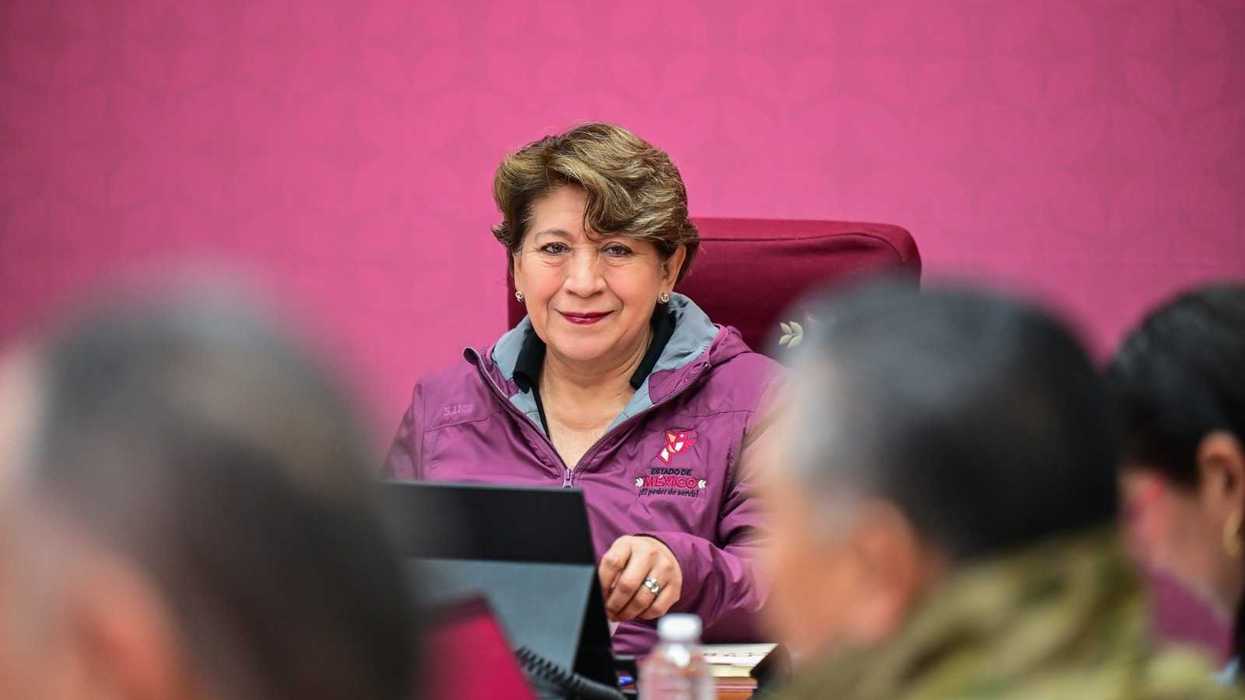 Woman in a magenta jacket smiles, seated at a table with others, against pink background.