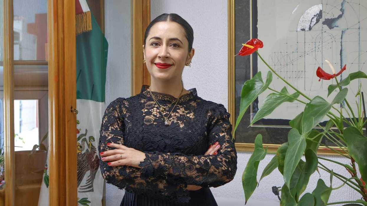 Woman in black lace dress, arms crossed, standing near Mexican flag and red flowers.