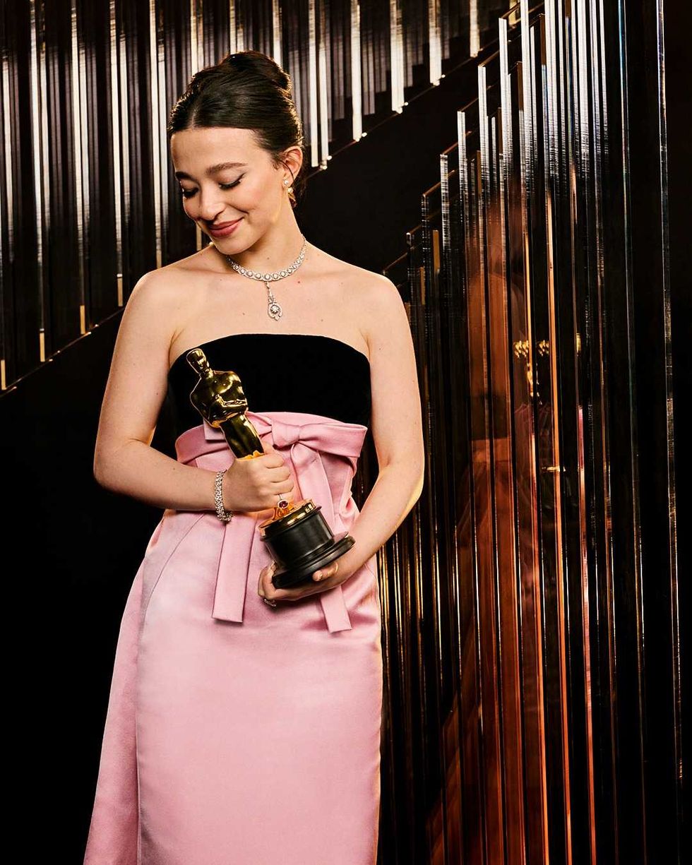 Woman in pink gown holding golden statuette, standing by reflective wall.