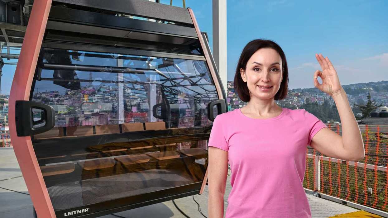 Woman in pink shirt gesturing "OK" beside a cable car with city view.