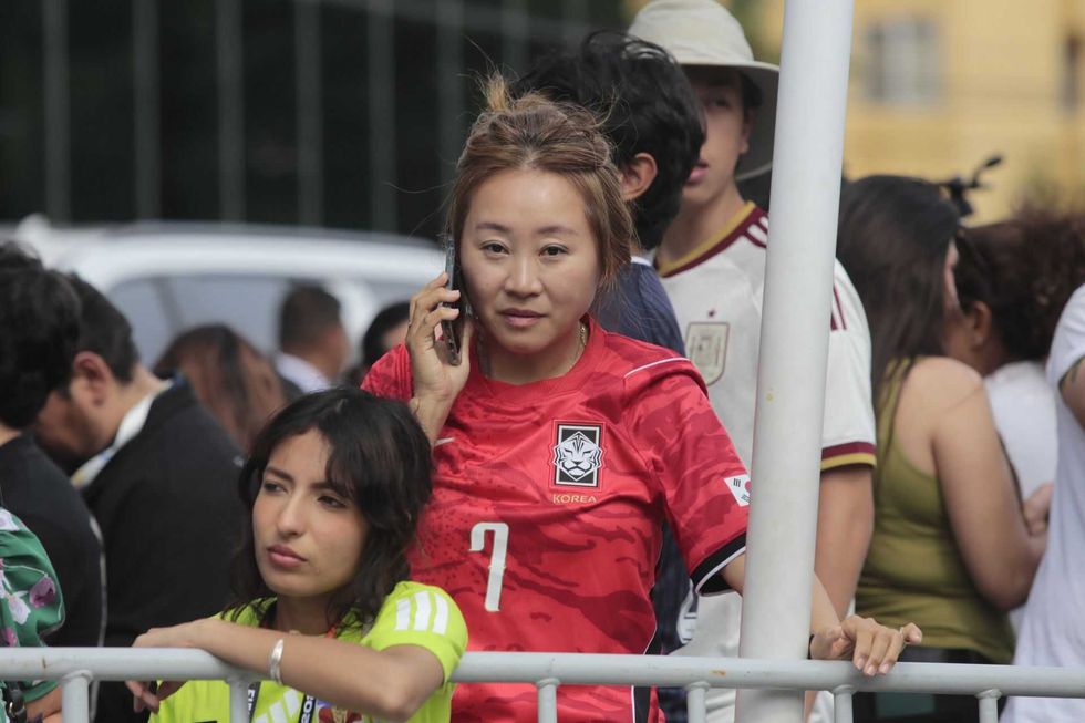 Woman in red Korea soccer jersey on phone at a crowded outdoor event.