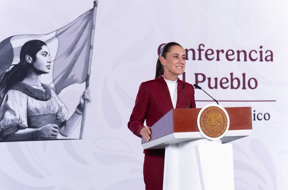 Woman in suit speaks at podium with historical mural in background.
