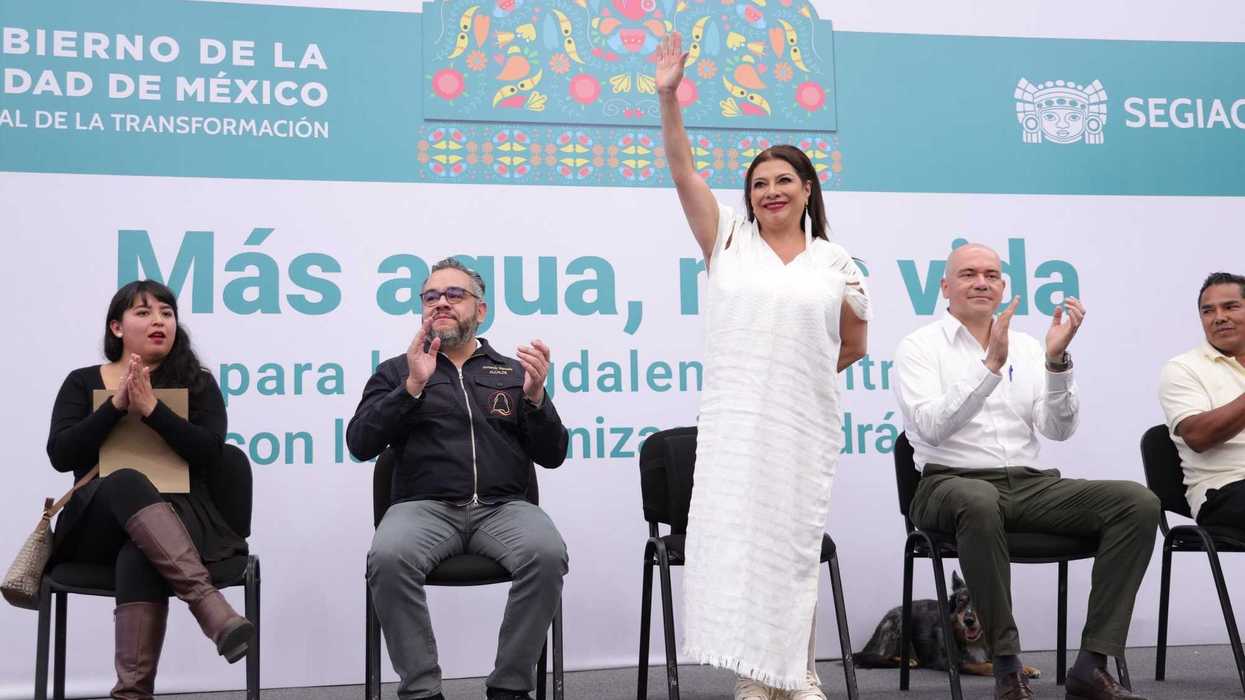 Woman in white dress waving, surrounded by seated people applauding at a public event.