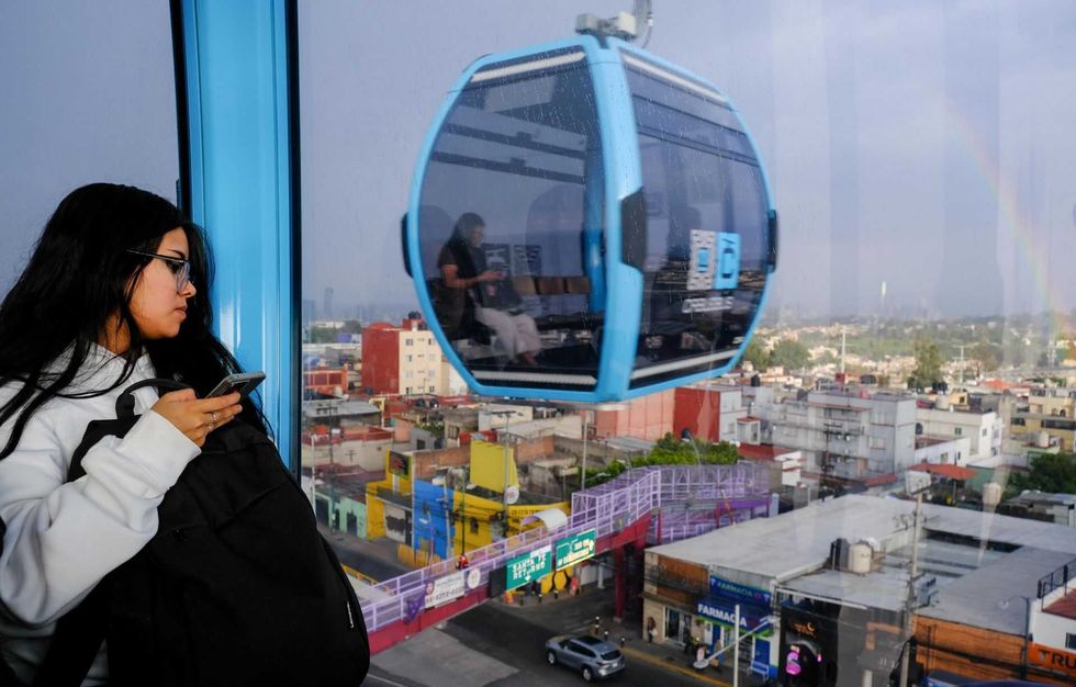 Woman on cable car in cityscape, holding phone; buildings and rainbow visible outside.