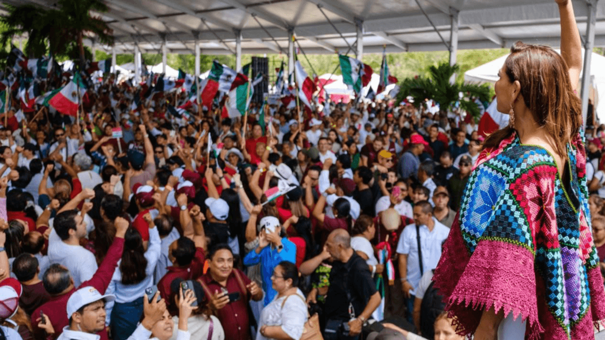 Woman raises fist at a large rally, surrounded by people waving flags under a canopy.
