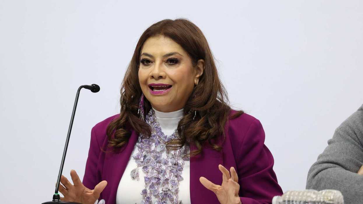 Woman speaking at a microphone, wearing a purple blazer and beaded necklace.