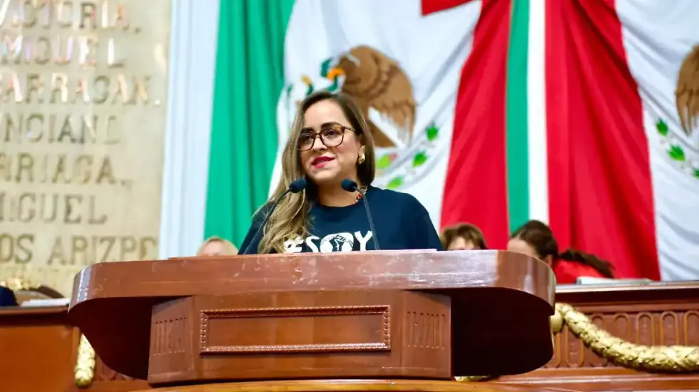 Woman speaking at a podium in front of a Mexican flag backdrop.