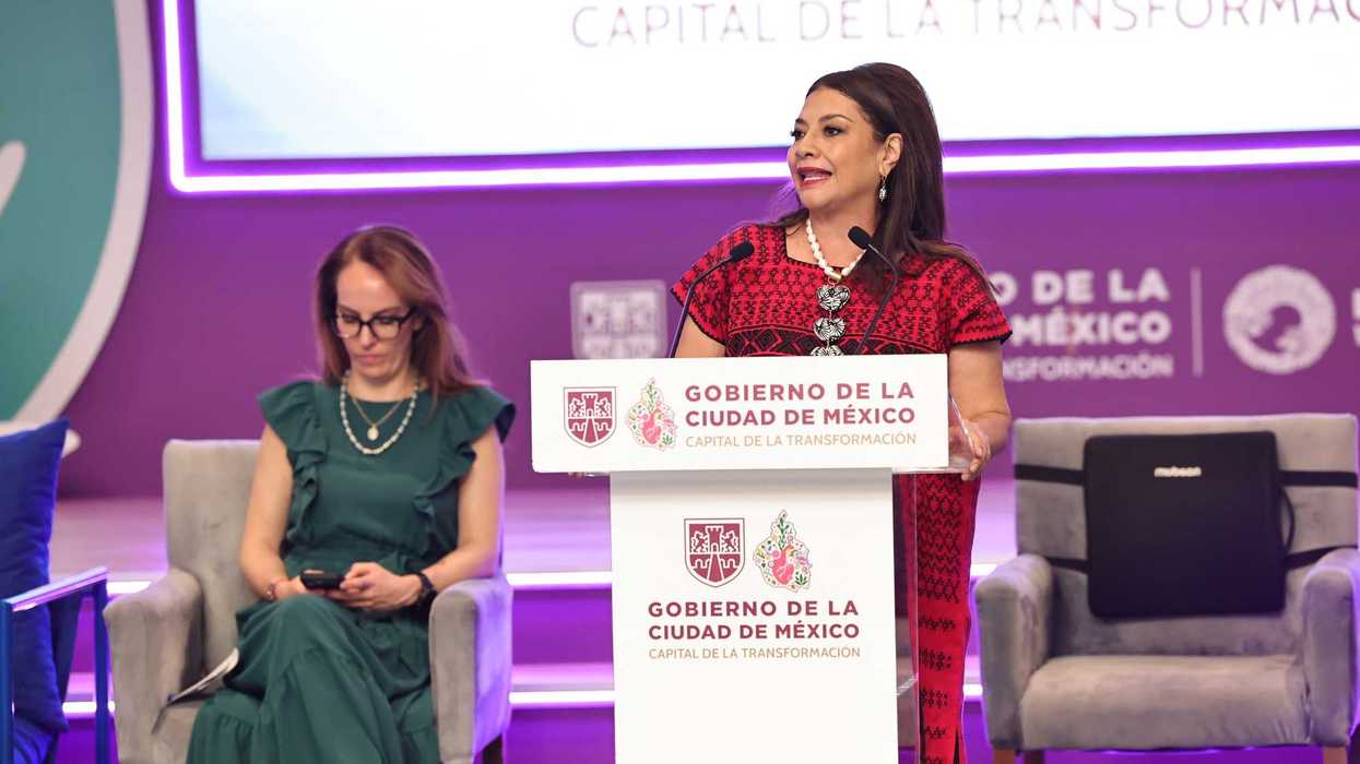 Woman speaking at a podium in Mexico City event, another woman seated nearby.