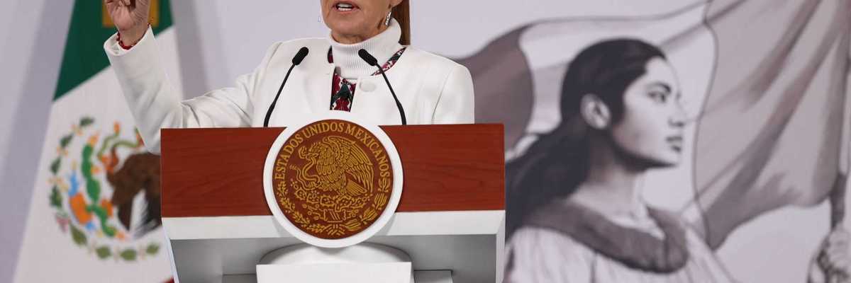 Woman speaking at a podium, Mexican flag and mural in the background.