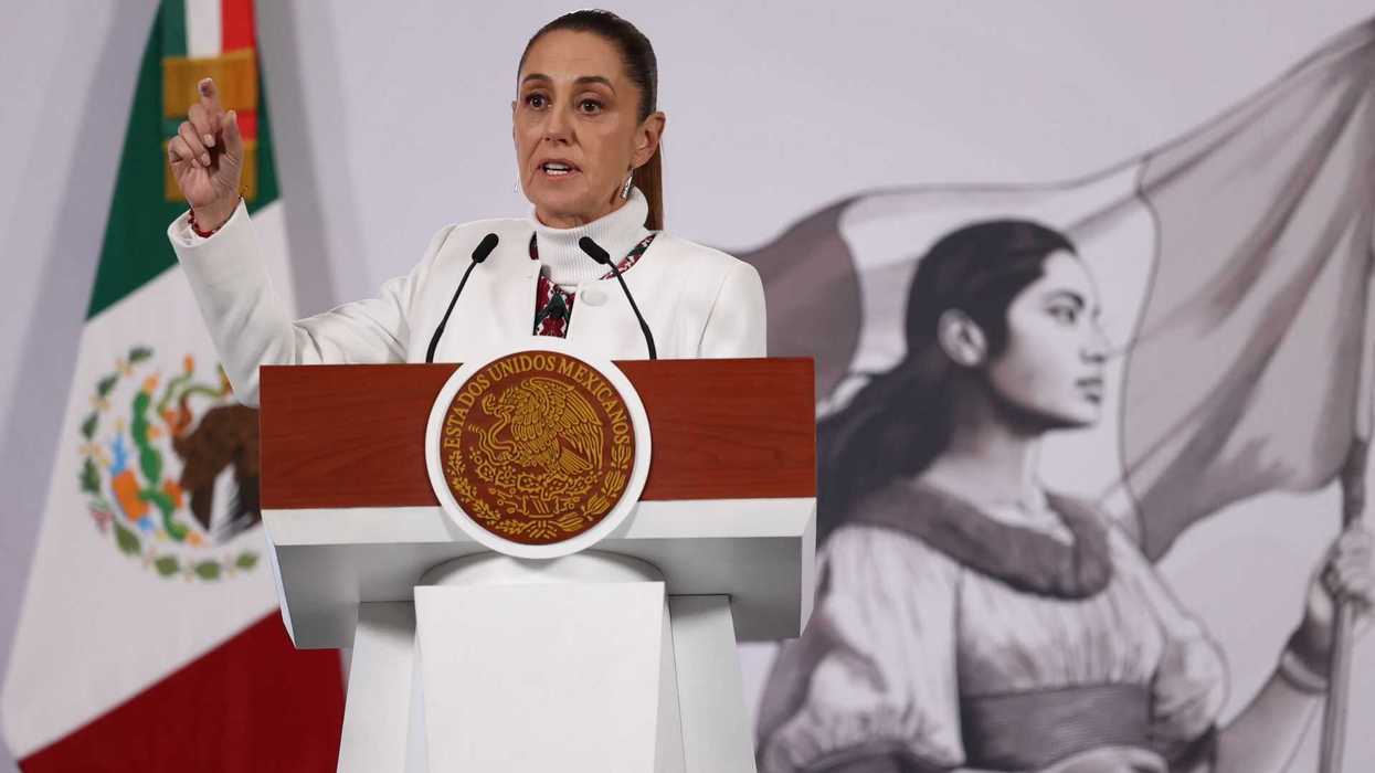 Woman speaking at a podium, Mexican flag and mural in the background.