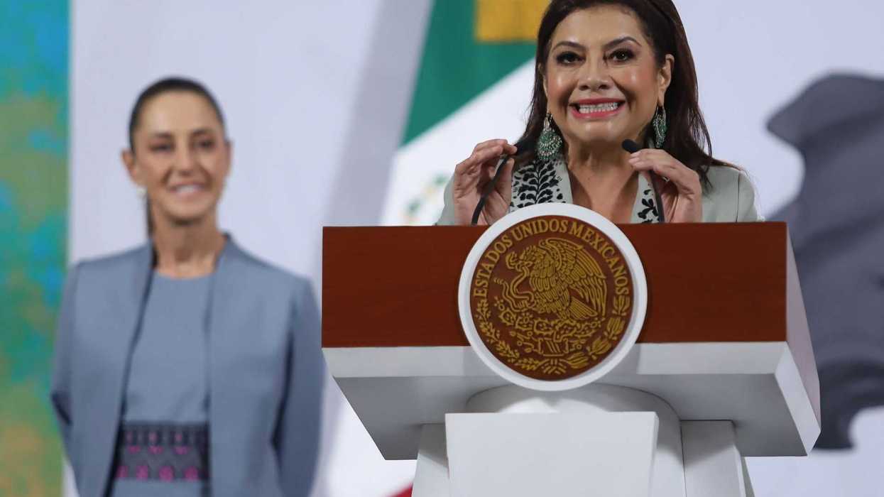 Woman speaking at a podium with Mexican insignia; another woman stands in the background, smiling.
