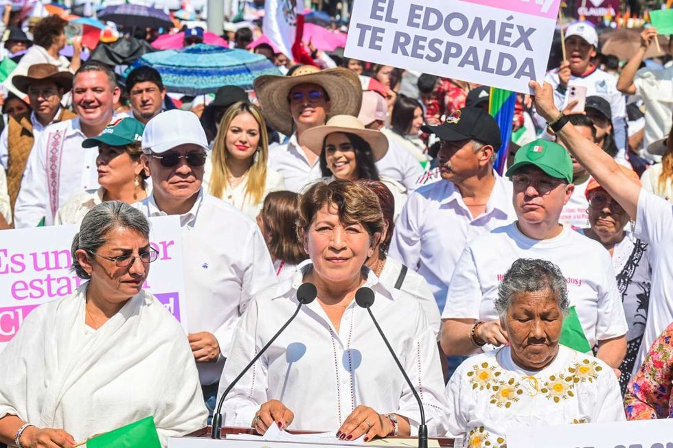 Woman speaking at a rally with a supportive crowd holding signs behind her.
