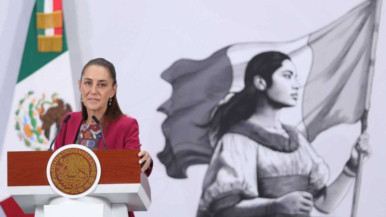 Woman speaking at podium, Mexican flag and mural of a woman with flag in background.