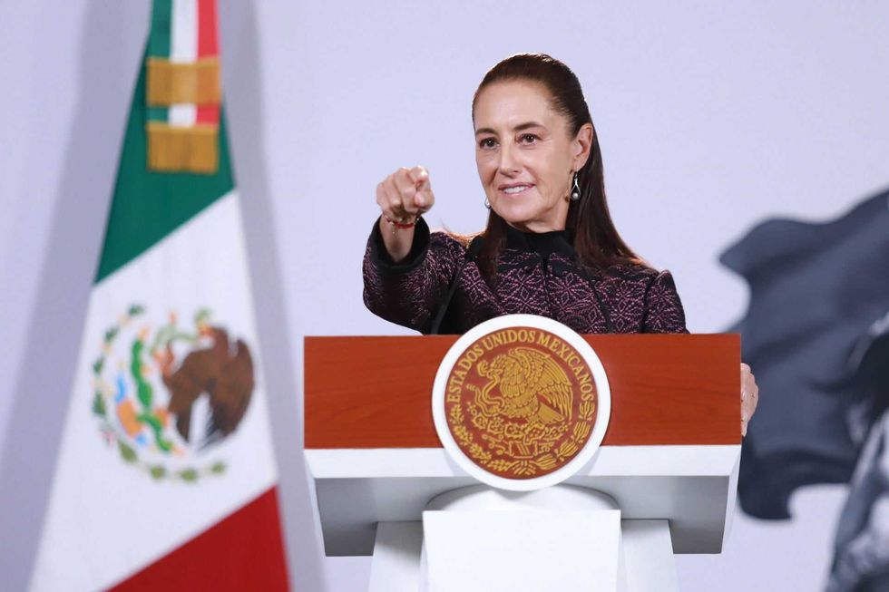 Woman speaking at podium with Mexican flag, pointing forward.