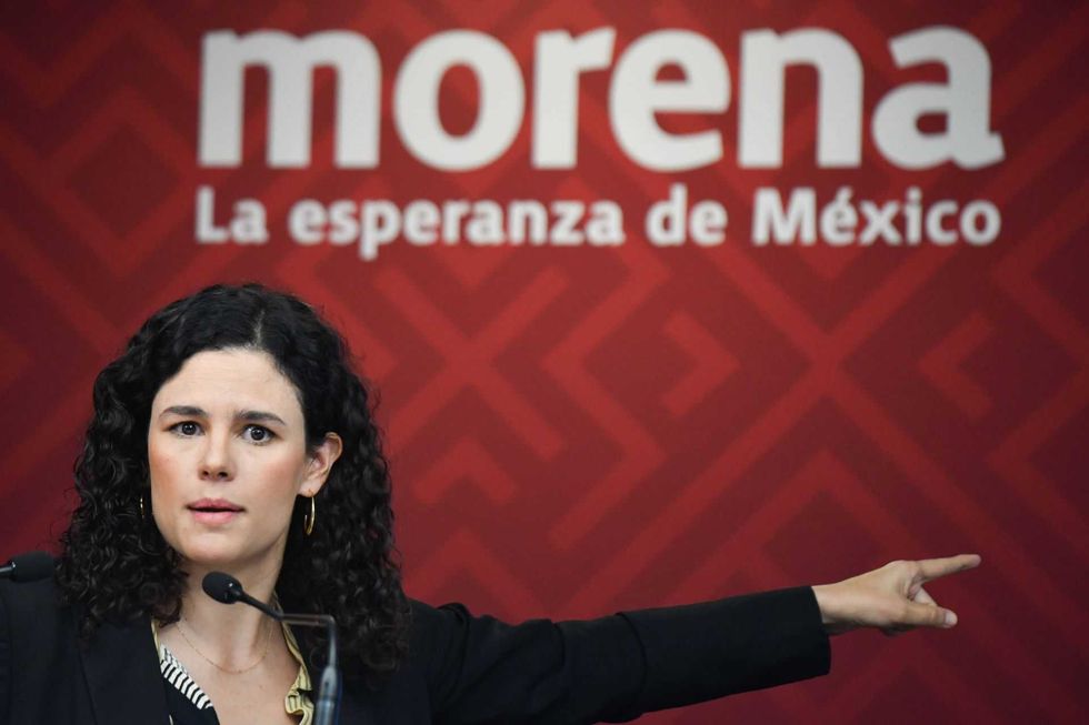 Woman speaking, pointing, in front of a "Morena: La esperanza de M\u00e9xico" sign.