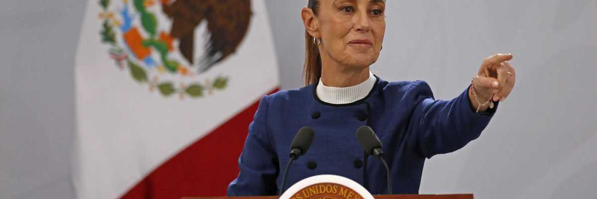 Woman speaks at podium with Mexican flag backdrop, gesturing forward.