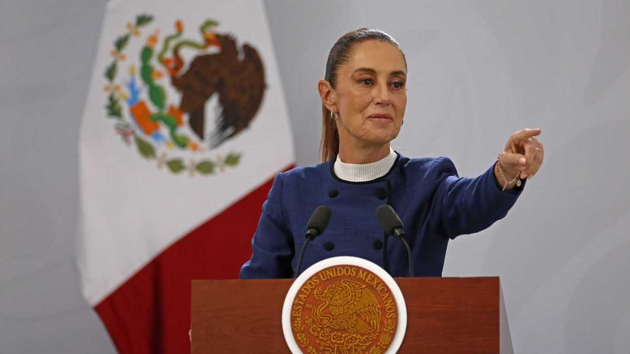 Woman speaks at podium with Mexican flag backdrop, gesturing forward.