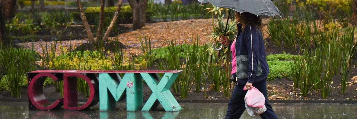 Woman walking in rain with umbrella by "CDMX" sculpture, wet pavement reflects image.