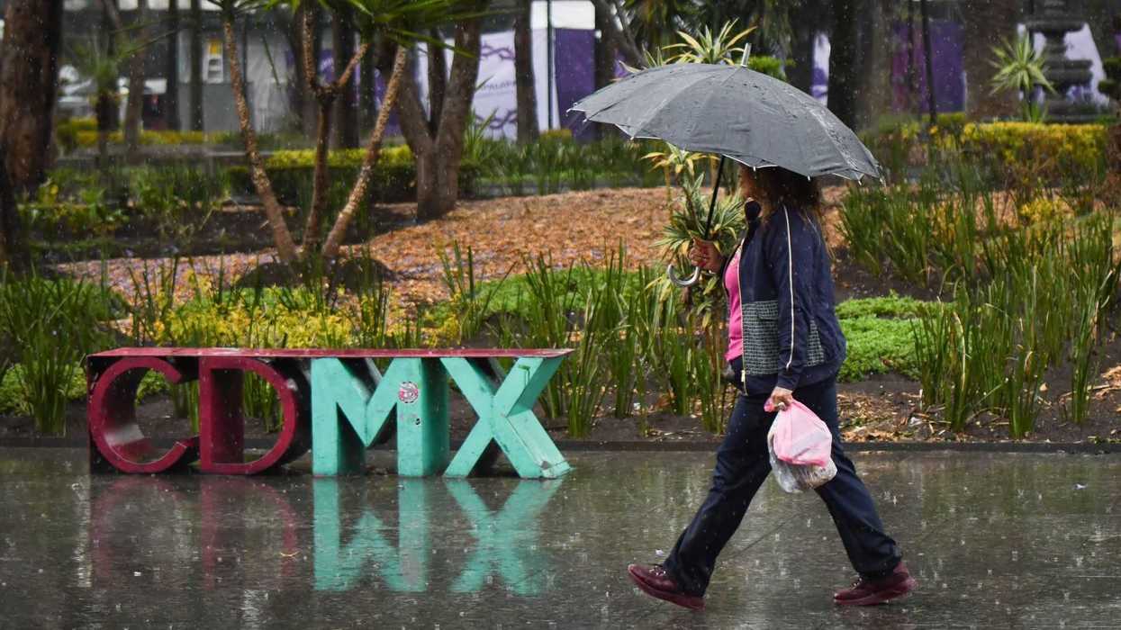 Woman walking in rain with umbrella by "CDMX" sculpture, wet pavement reflects image.