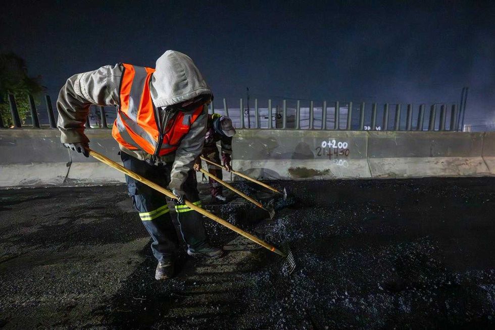 Workers with rakes lay asphalt on a road at night, wearing reflective gear.