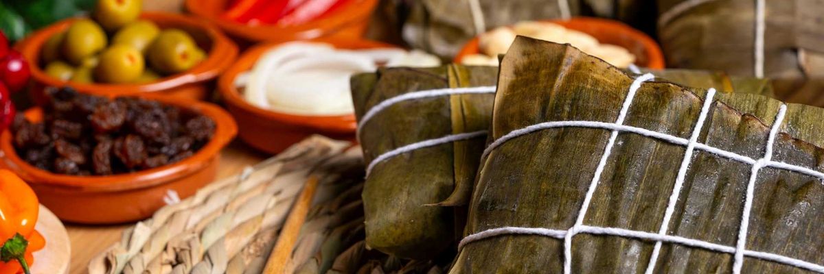 Wrapped tamales on a woven mat, with olives, peppers, and other ingredients in bowls.